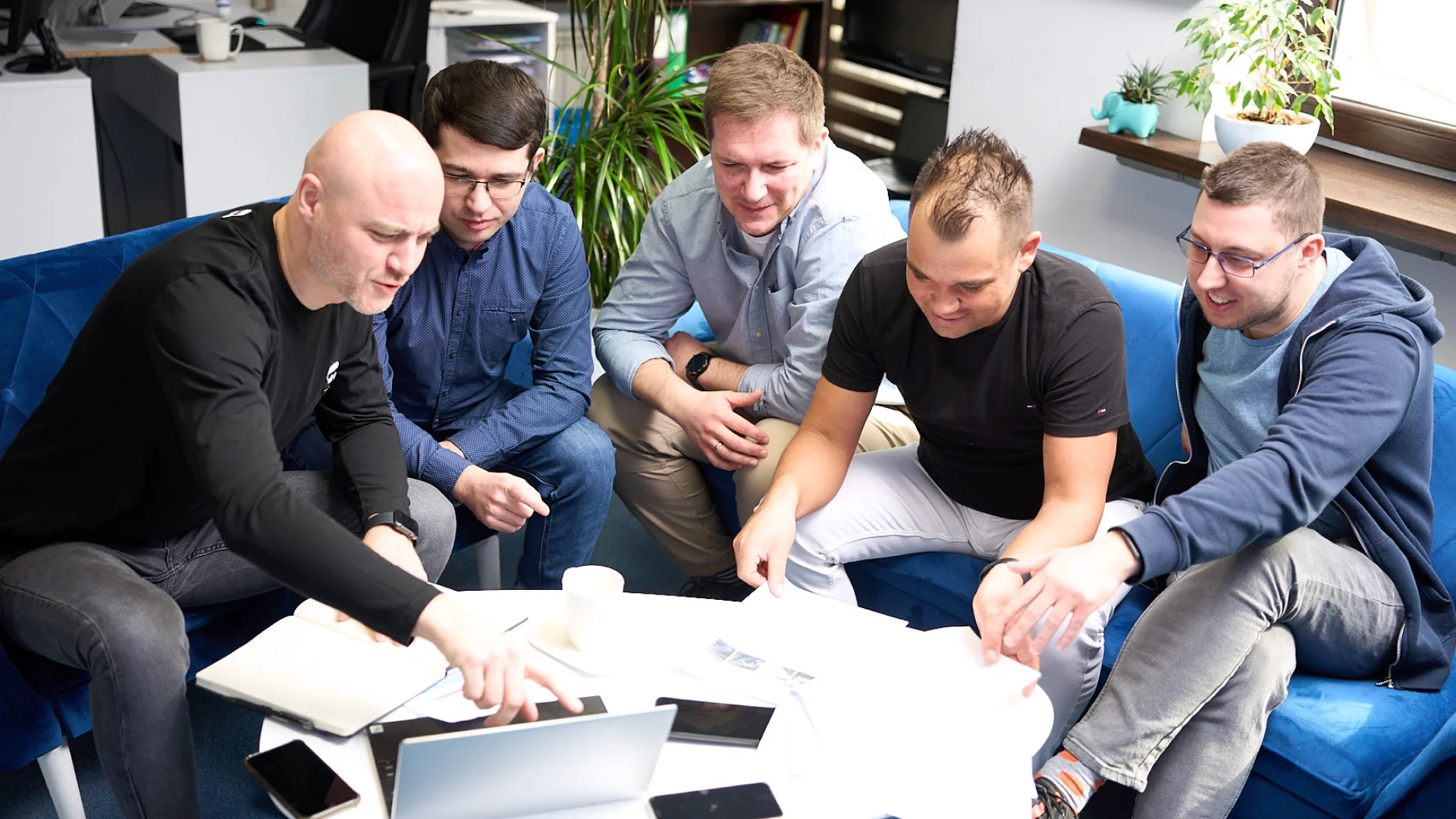 Zdjęcie przedstawia grupę mężczyzn w biurze, omawiających projekt. Na pierwszym planie tekst promocyjny firmy zajmującej się marketingiem.